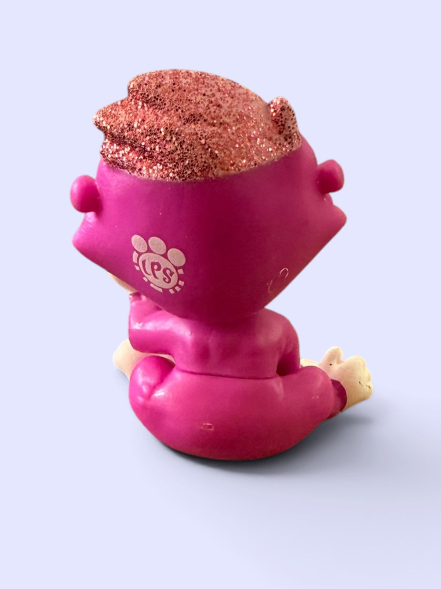 A small pink figurine or toy shown from the back, with glittery copper-brown hair and a white paw-print style logo on the back, photographed against a pale lavender background.