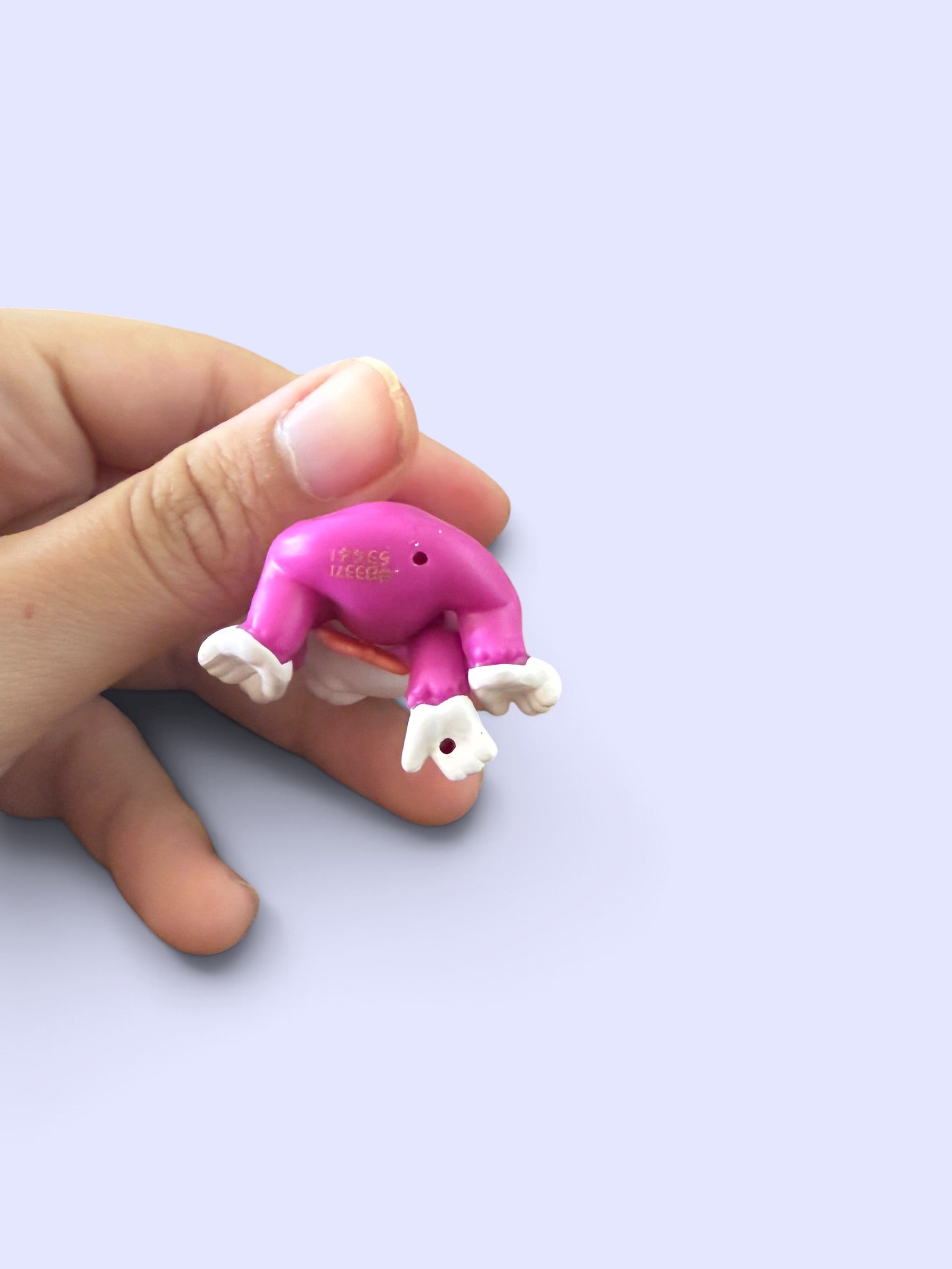 A small pink and white plastic toy figure shaped like a bent figure, held between a person's fingers against a light lavender background.