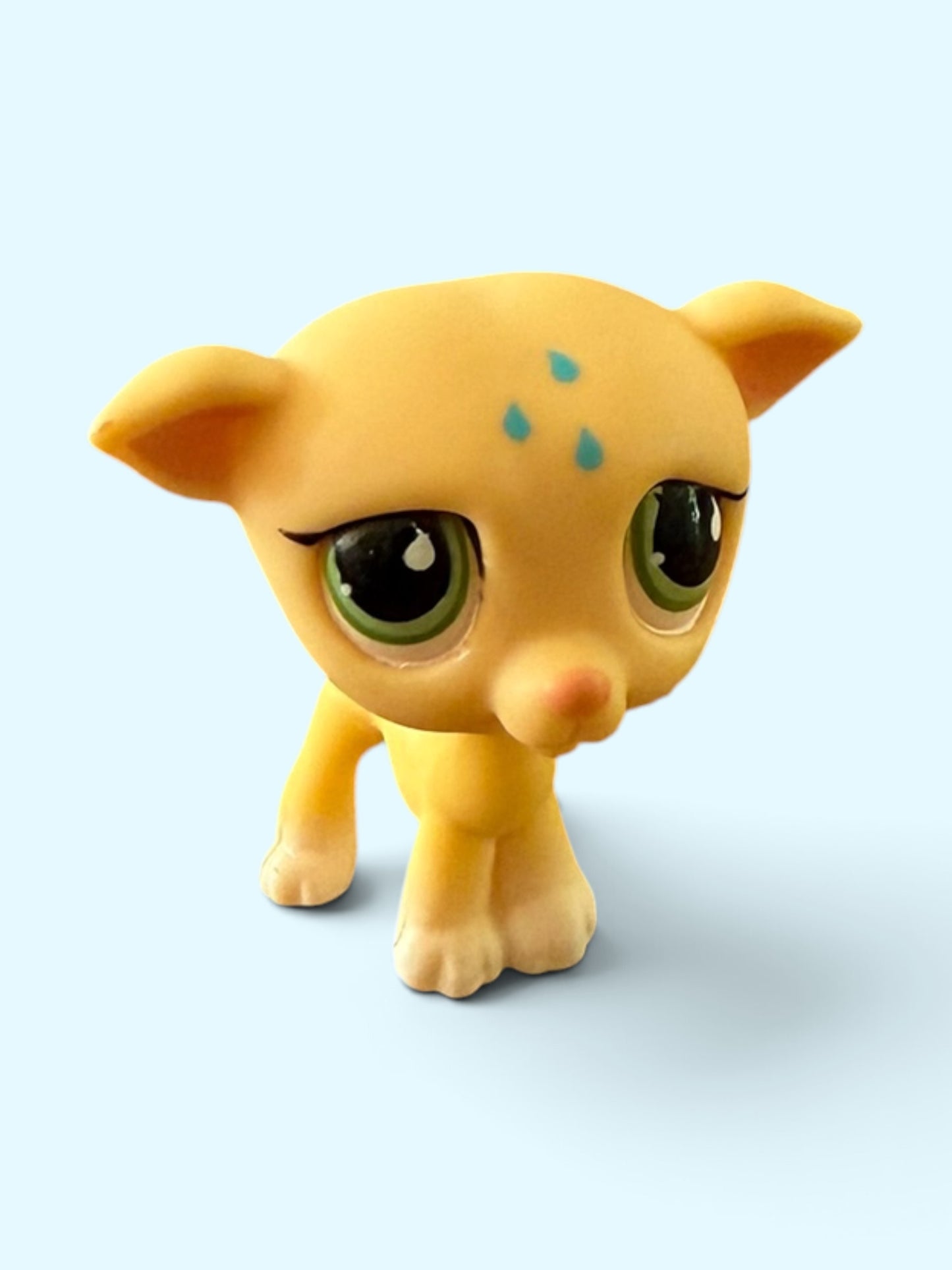 A small yellow toy animal figure with large green eyes, black eyelashes, and three small blue spots on its forehead, standing against a light blue background.