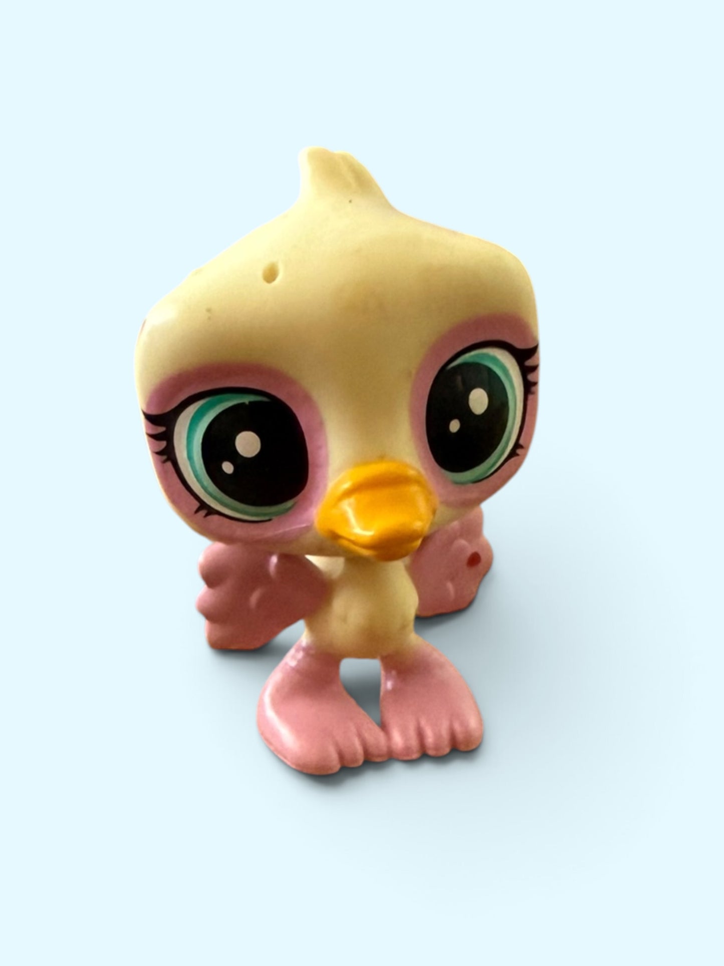 A small plastic toy duckling figure with oversized cartoon eyes, a yellow-orange beak, and pink feet and wing details, shown against a light blue background.