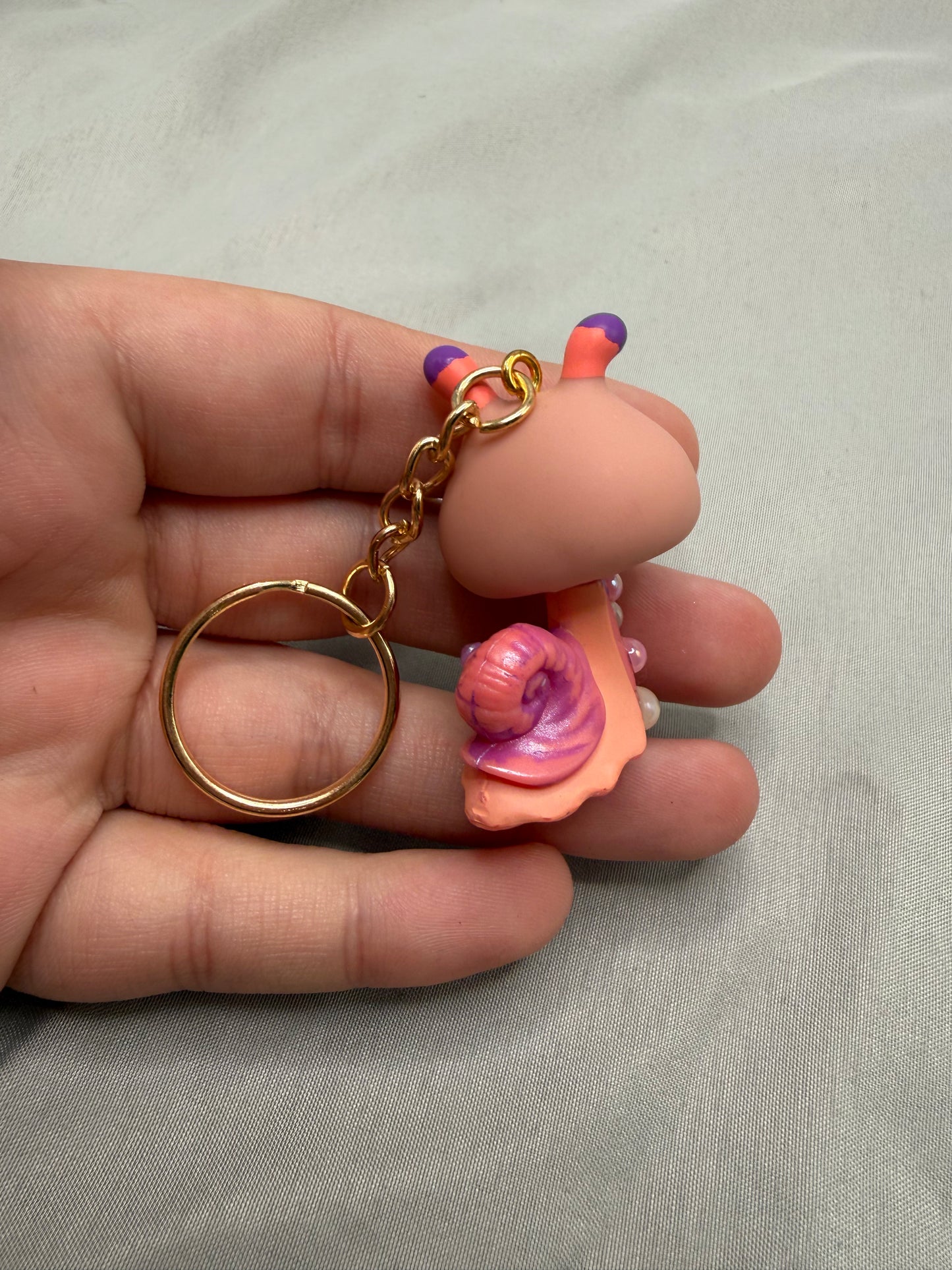 Hand holding a keychain with a pink shell and gold ring on a light gray background