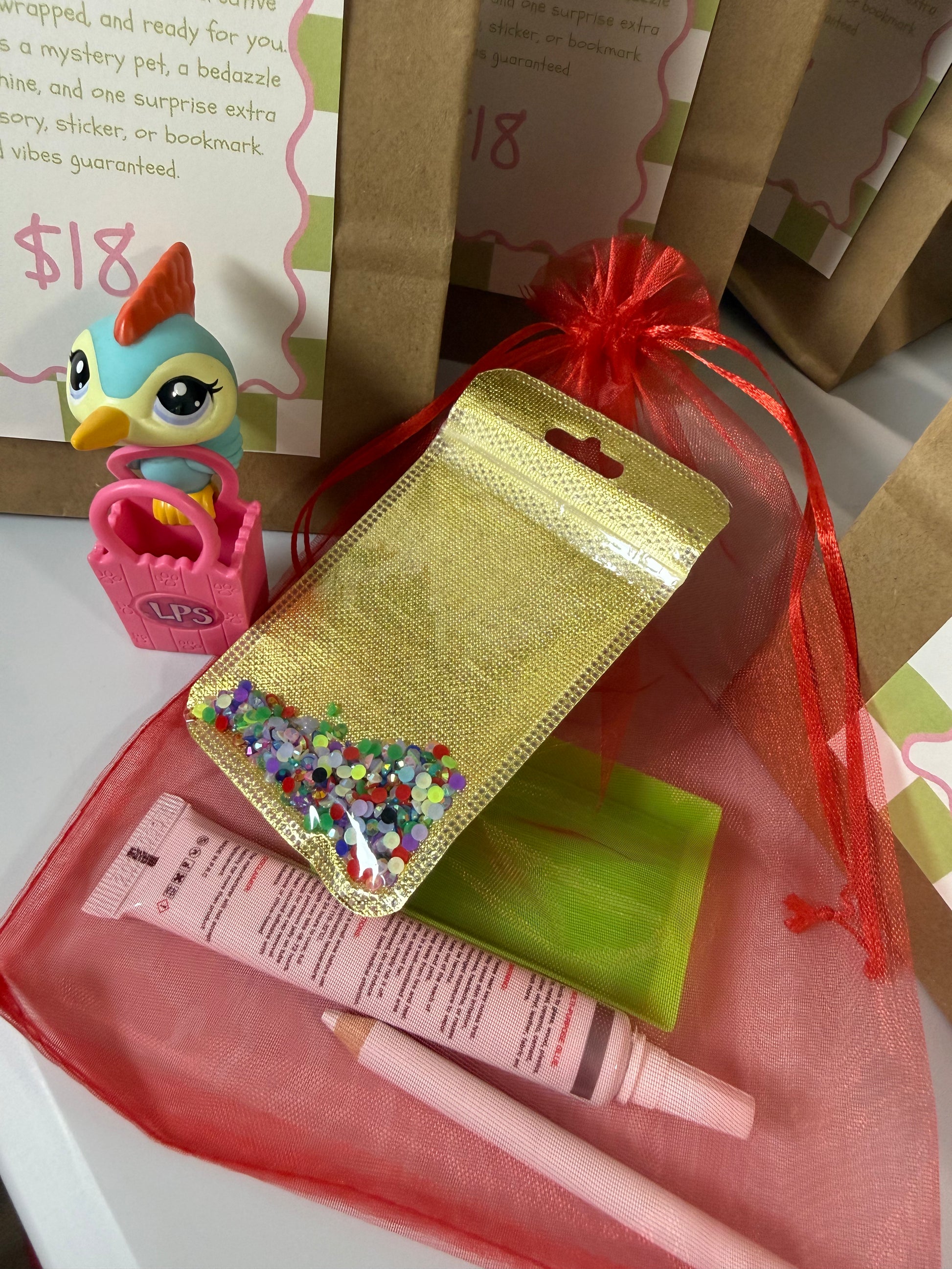 A mystery pet surprise box set displayed on a white surface, featuring a small blue bird figurine in a pink basket, a gold glitter pouch filled with colorful confetti, a pink makeup tube, a pink pencil, a green square item, and red sheer organza bags.