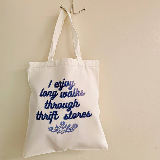 White tote bag with blue text and floral design hanging on a hook against a beige wall.
