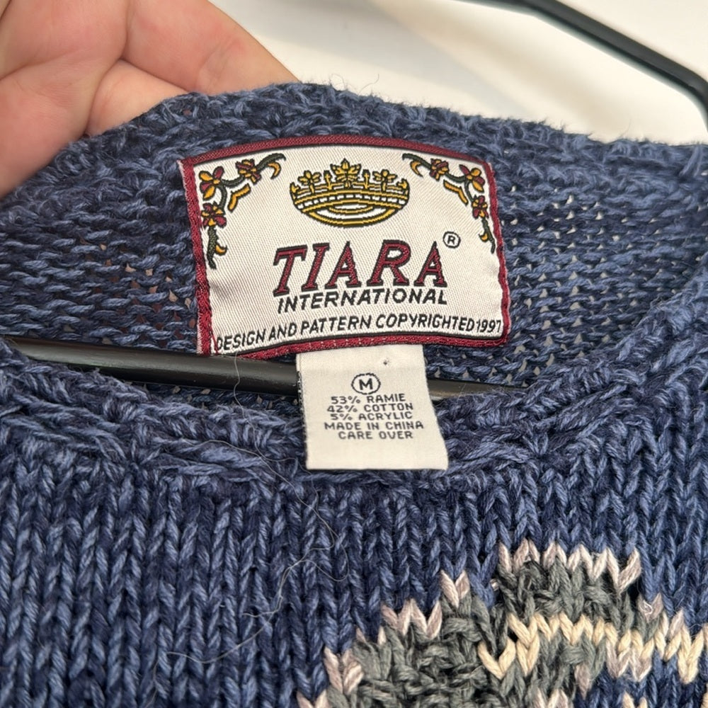 Closeup of Tiara International Tag of Blue knit sweater with an allover floral and botanical pattern, long sleeves, and ribbed hem.