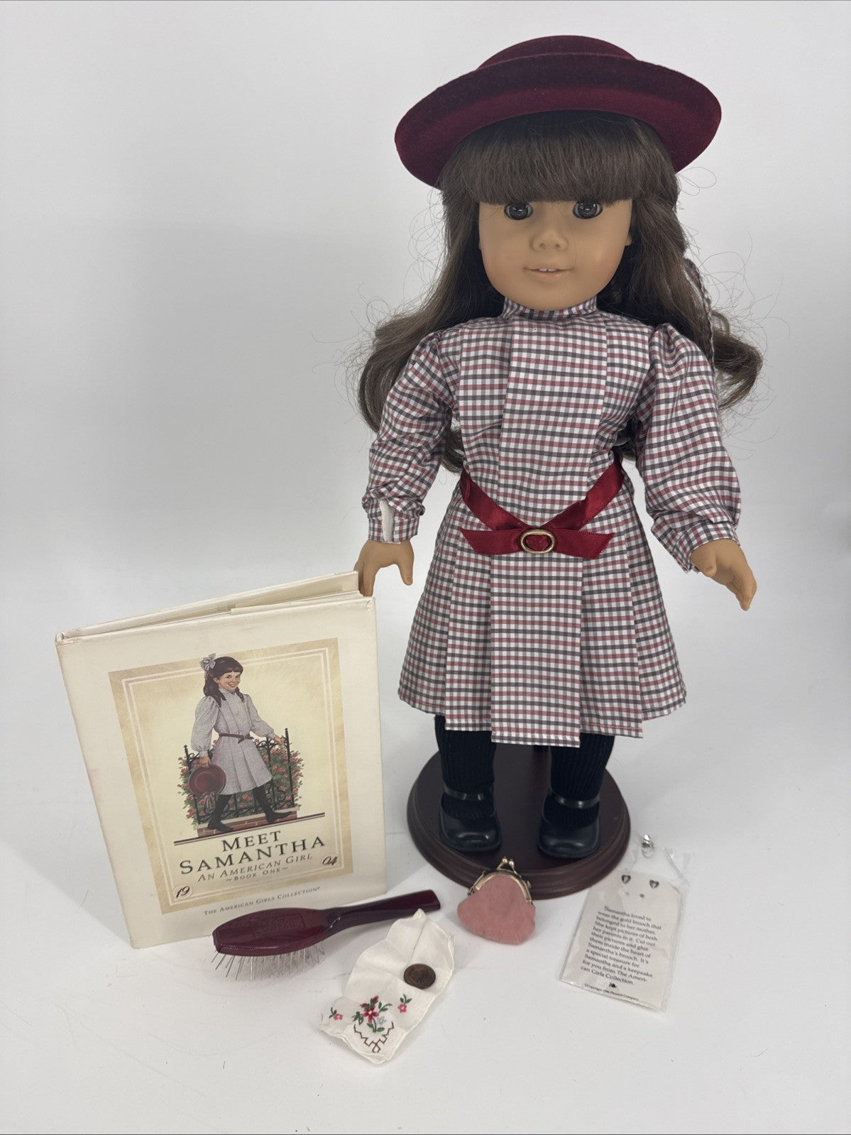 Doll with checkered dress and red hat, accompanied by a book and accessories on a white background