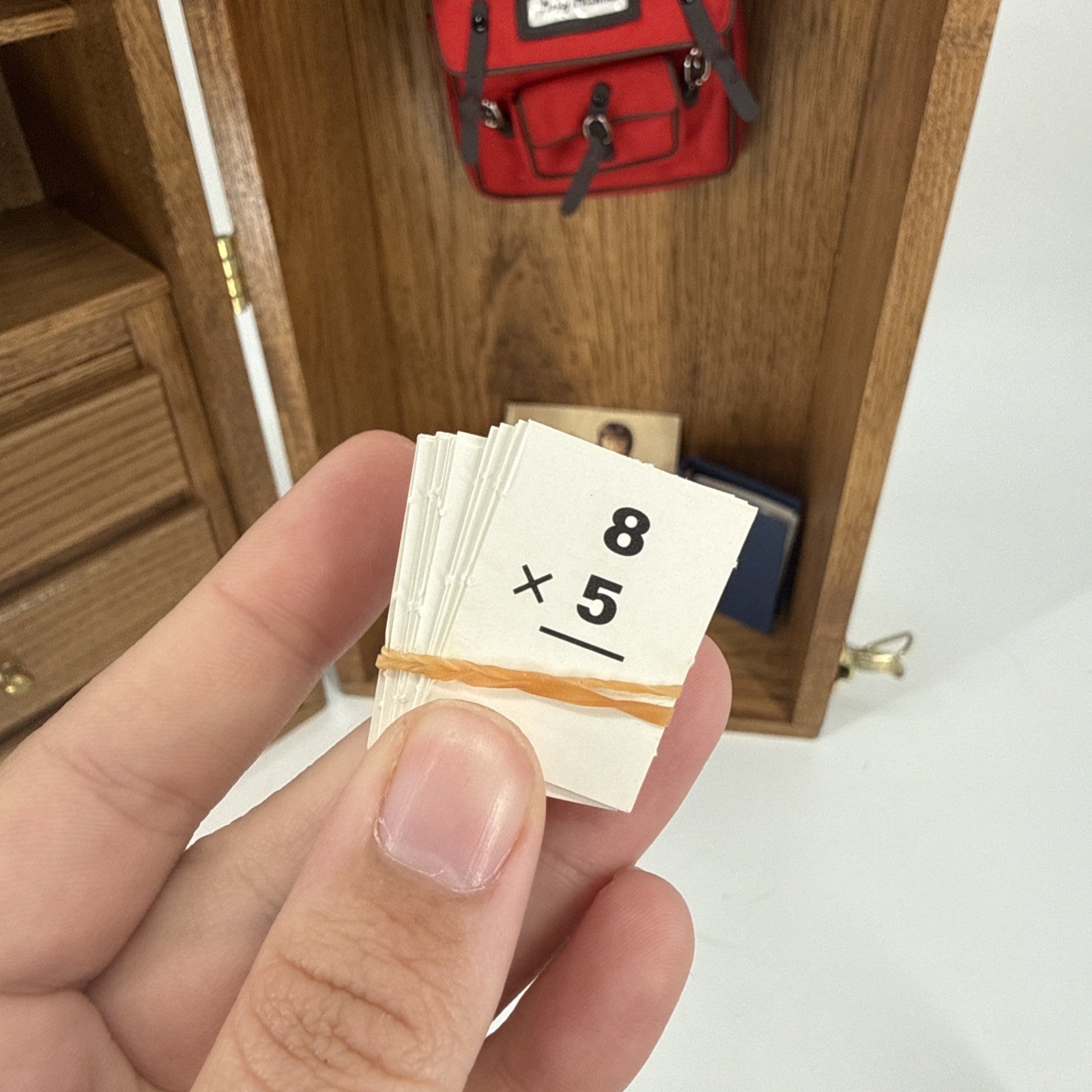 Hand holding a small stack of paper with multiplication problem, red backpack in background
