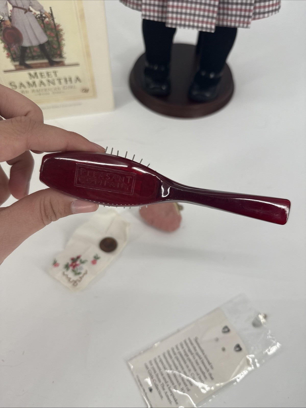 Red hairbrush held by a hand on a white surface with a book and small figurine in the background.