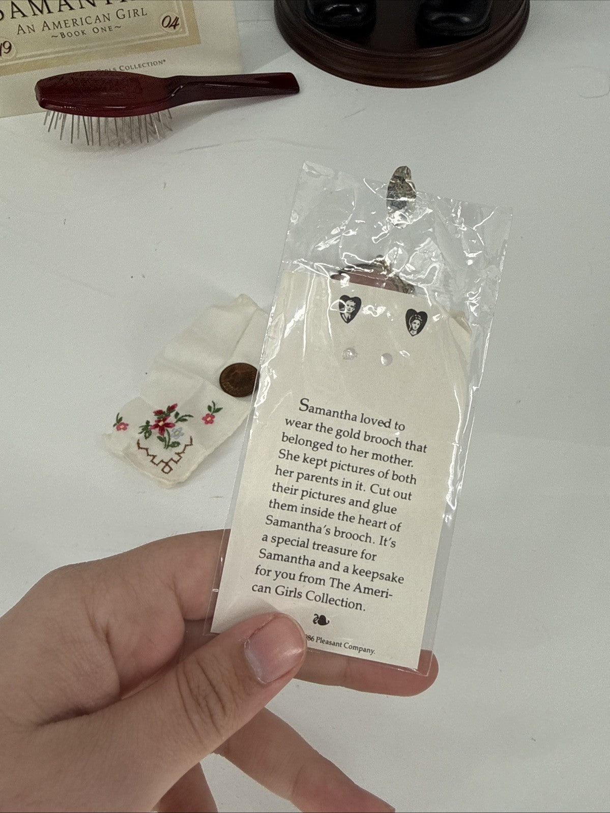 Hand holding a tag with text and an American Girl branded brooch on a white surface.