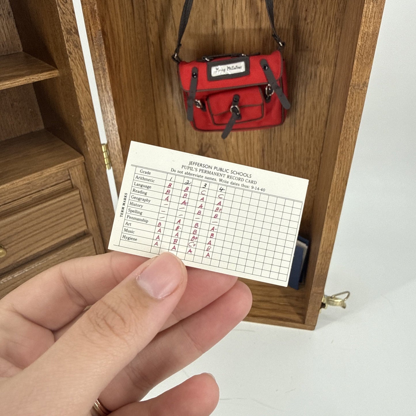 Hand holding a miniature record card in front of a wooden cabinet with a red handbag on top.