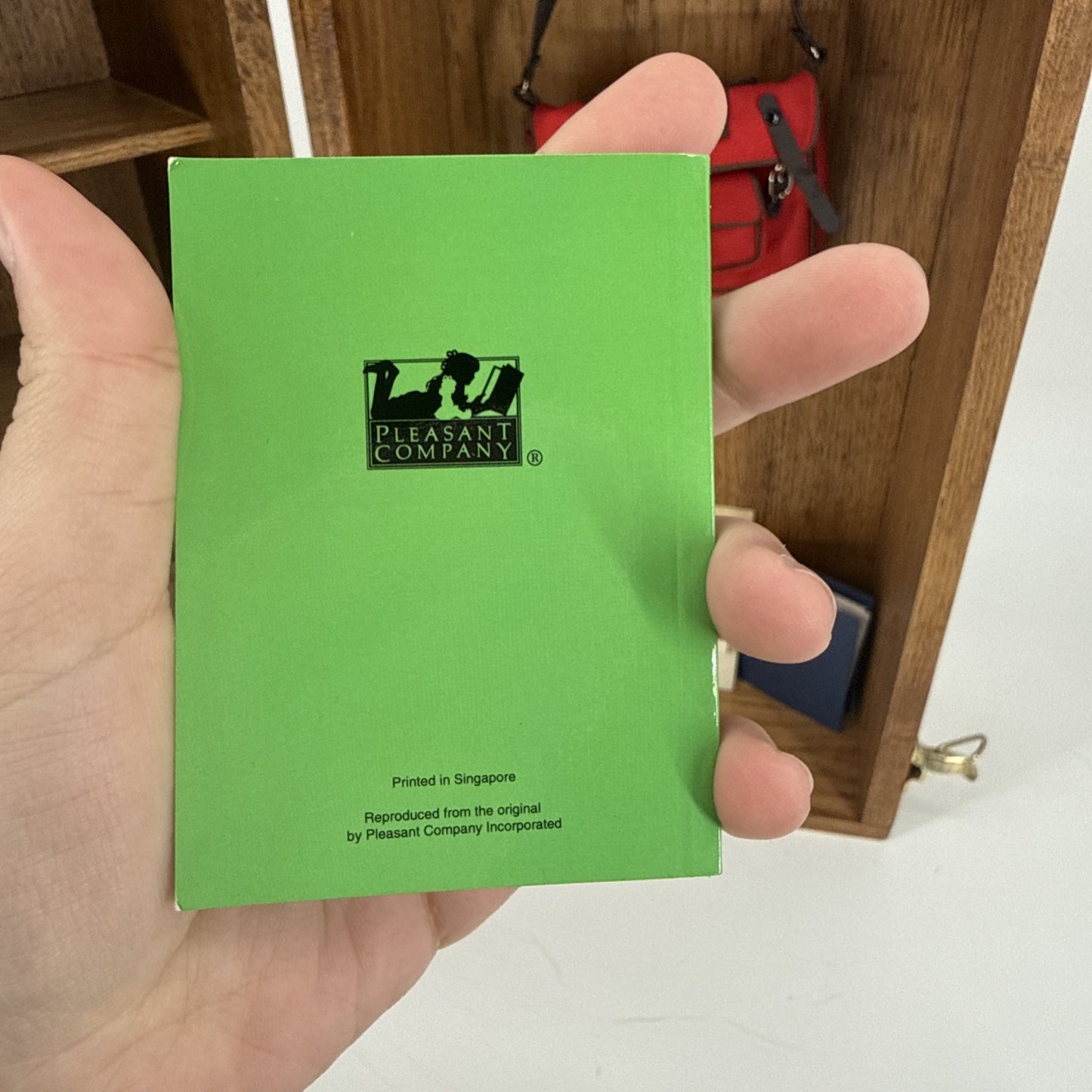 Hand holding a green card with Pleasant Company logo against a wooden shelf background