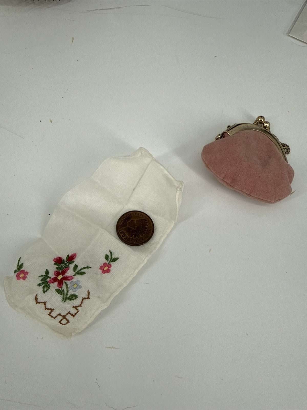 Small embroidered fabric piece with floral designs and a coin, next to a pink fabric pouch on a white surface.